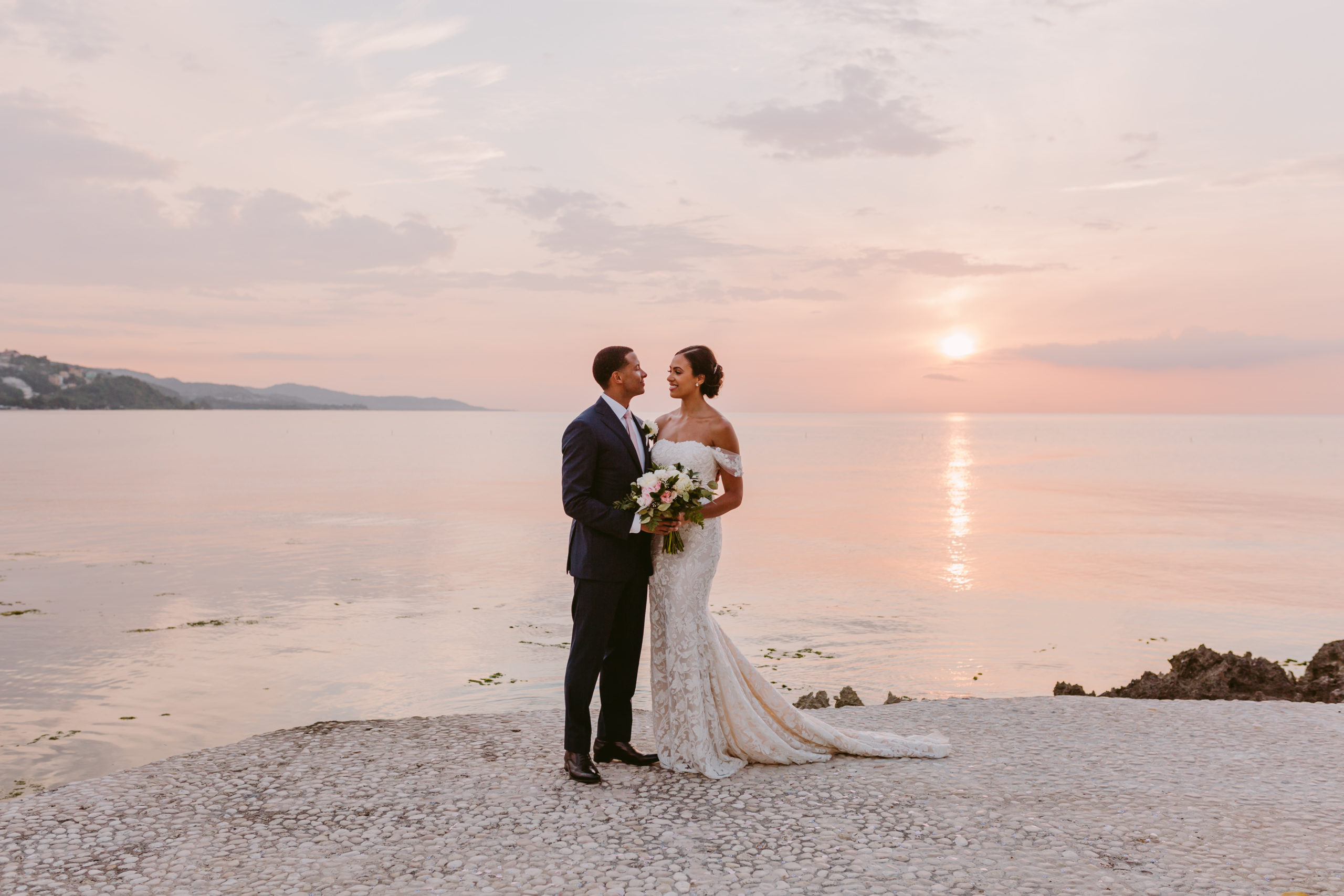 Errol Barnett & Ariana Tolbert stand on the shore of Montego Bay smiling at each other. Image by Victoria Heer, fine art wedding photographer.